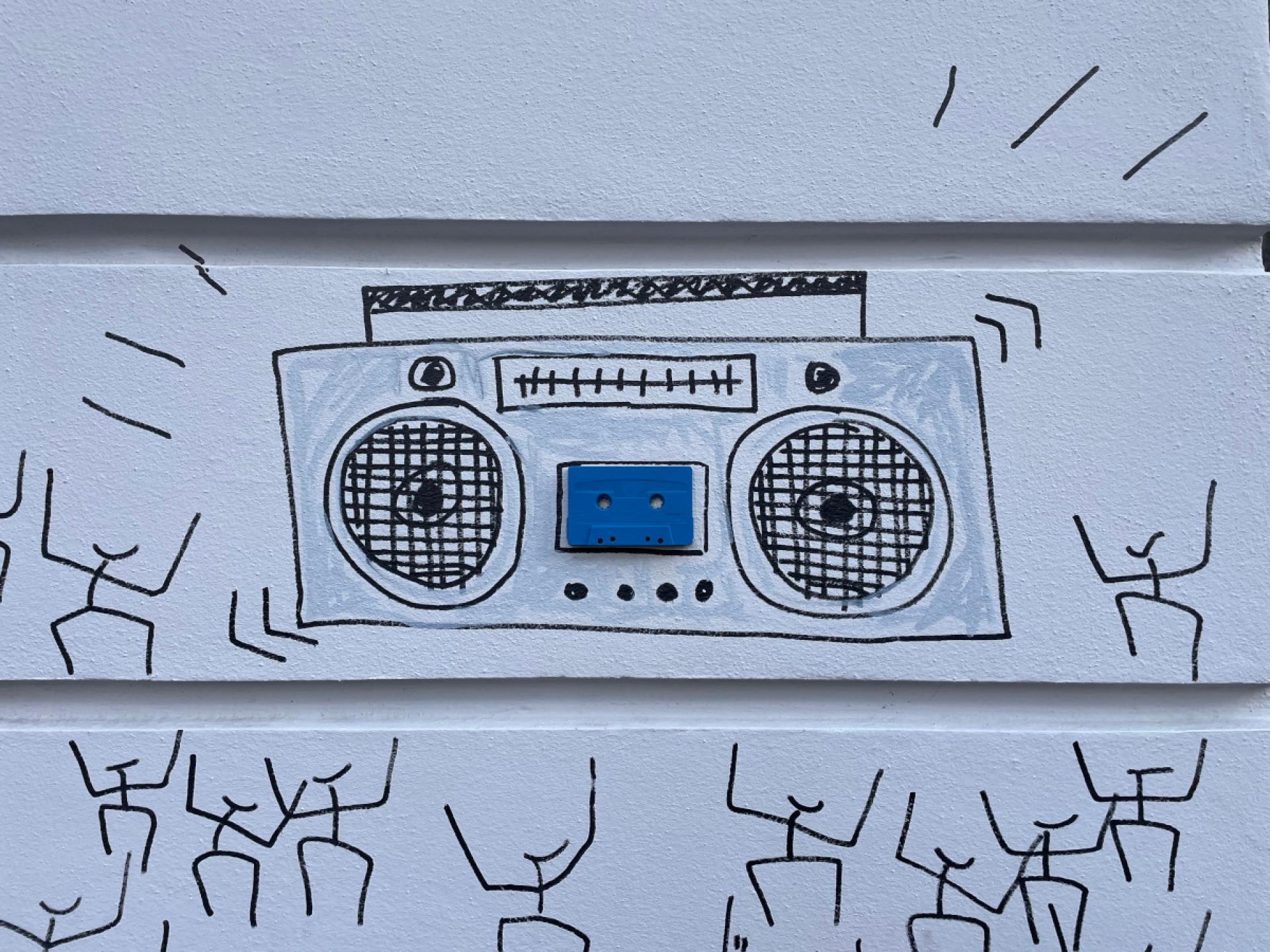 A cassette tape stuck to the wall of a house. Someone has drawn a boombox and dancing stick figures around it.