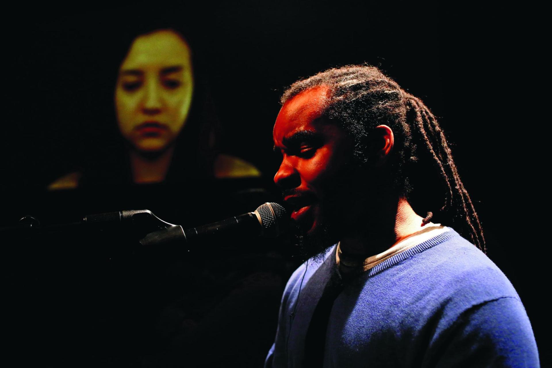 Eine Person of Color singt im Vordergrund in ein Mikrofon und im Hintergrund sieht man auf einem großen Bildschirm ein Gesicht.
