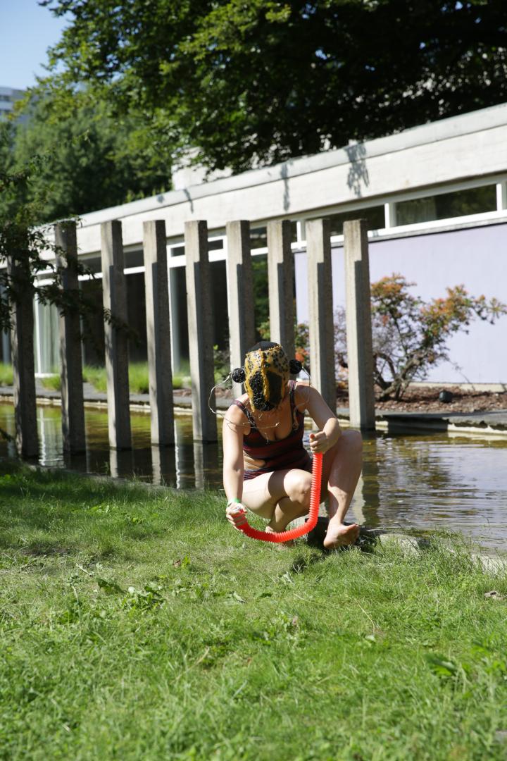 Performance draussen im Hof der Akademie der Künste, die Performerin trägt Badeanzug und Maske und hockt auf dem Rasen