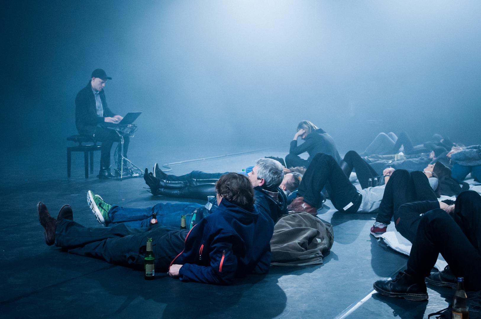 Links der Performer auf einem Stuhl, rechts von ihm auf dem Boden liegende Besucher:innen