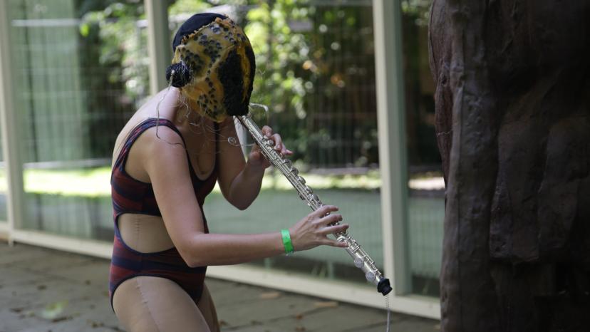 Eine Klangperformance mit einer Person draussen im Hof der Akademie der Künste, die Person trägt Badeanzug und eine Maske und spielt ein Instrument